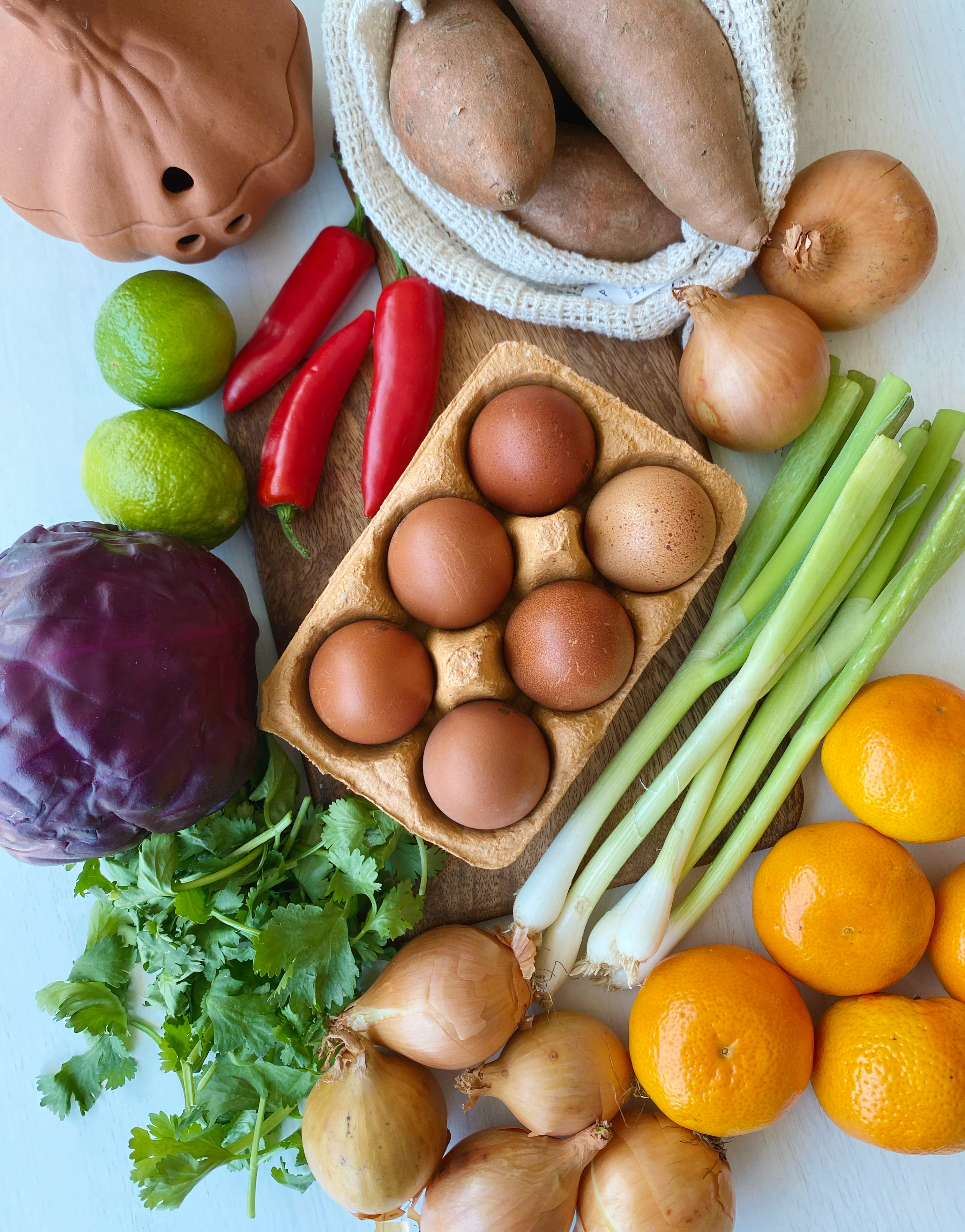 A collection of healthy foods including eggs, oranges, onions, sweet potatoes, chillis, red cabbage, coriander and limes.