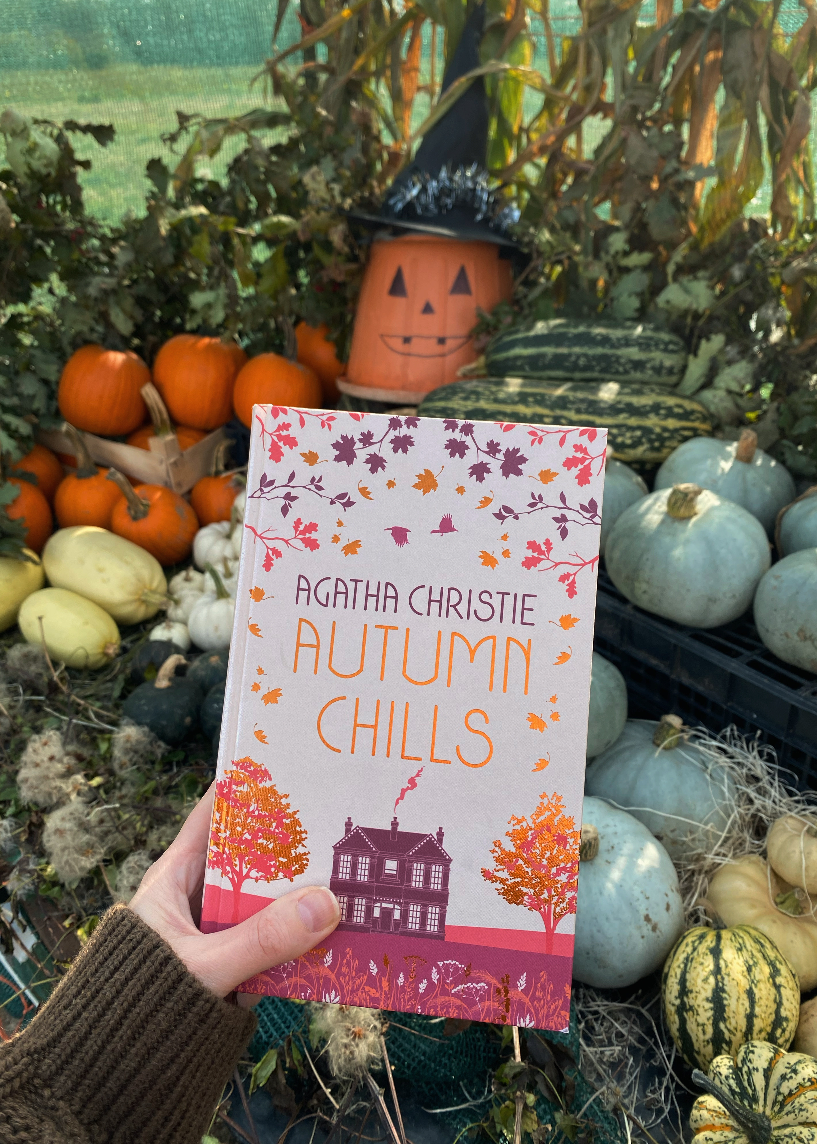 A hand in a brown jumper holding a copy of Agatha Christie's Autumn Chills book in front of an autumn Halloween pumpkin display.